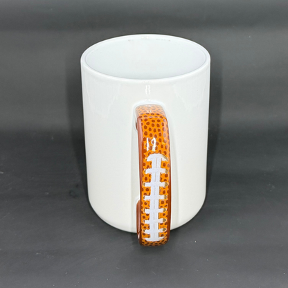 A glossy white ceramic mug with a handle shaped and painted like an American football.