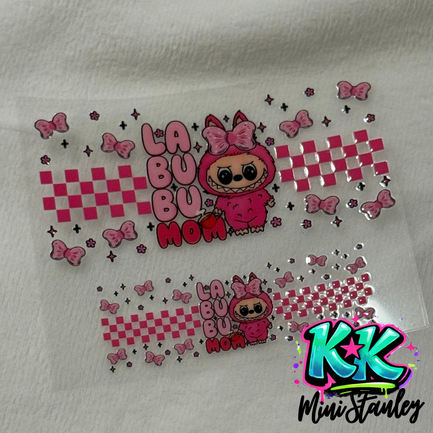 Two pink Labubu character UV DTF cup wraps with checkered patterns and bows on a clear sheet.