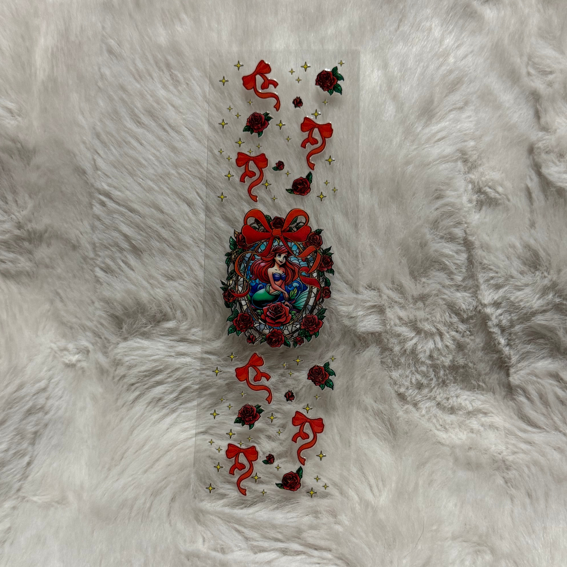 A clear UV DTF pen wrap featuring Ariel surrounded by red roses, bows, and gold stars.