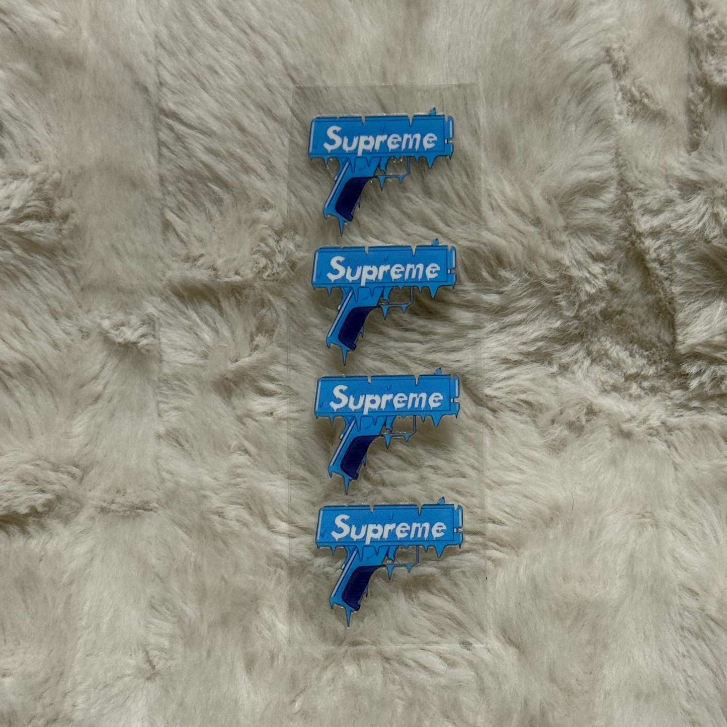 A sheet of four blue Supreme gun-themed UV DTF adhesive decals on a clear backing.