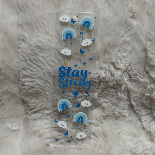 A clear adhesive pen wrap featuring blue rainbows, smiling clouds, butterflies, and Stay Strong text.
